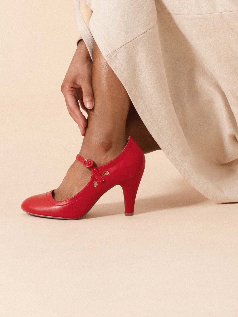 A hand fastening the strap of a red J.Adams Pixie Mary Jane heel, worn with a long cream skirt