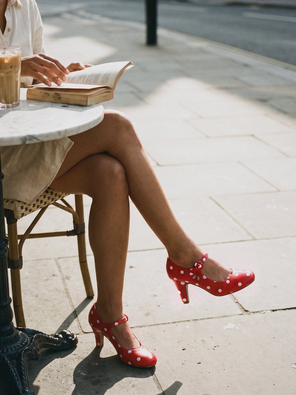 Red and white polka dot J.Adams Pixie Mary Jane heels on crossed legs at a Paris café table with an iced coffee and a book