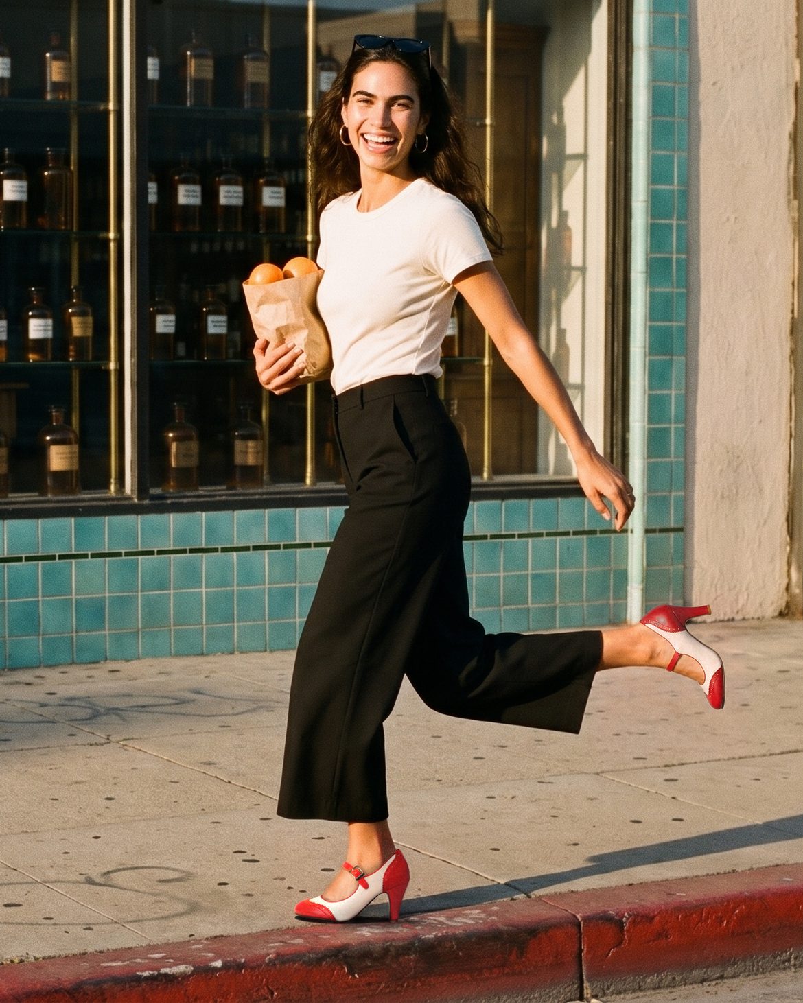 Smiling woman skipping along a sunny Los Angeles sidewalk past an apothecary window, holding a bag of oranges, wearing red-and-cream J.Adams Kym wingtip Mary Jane heels
